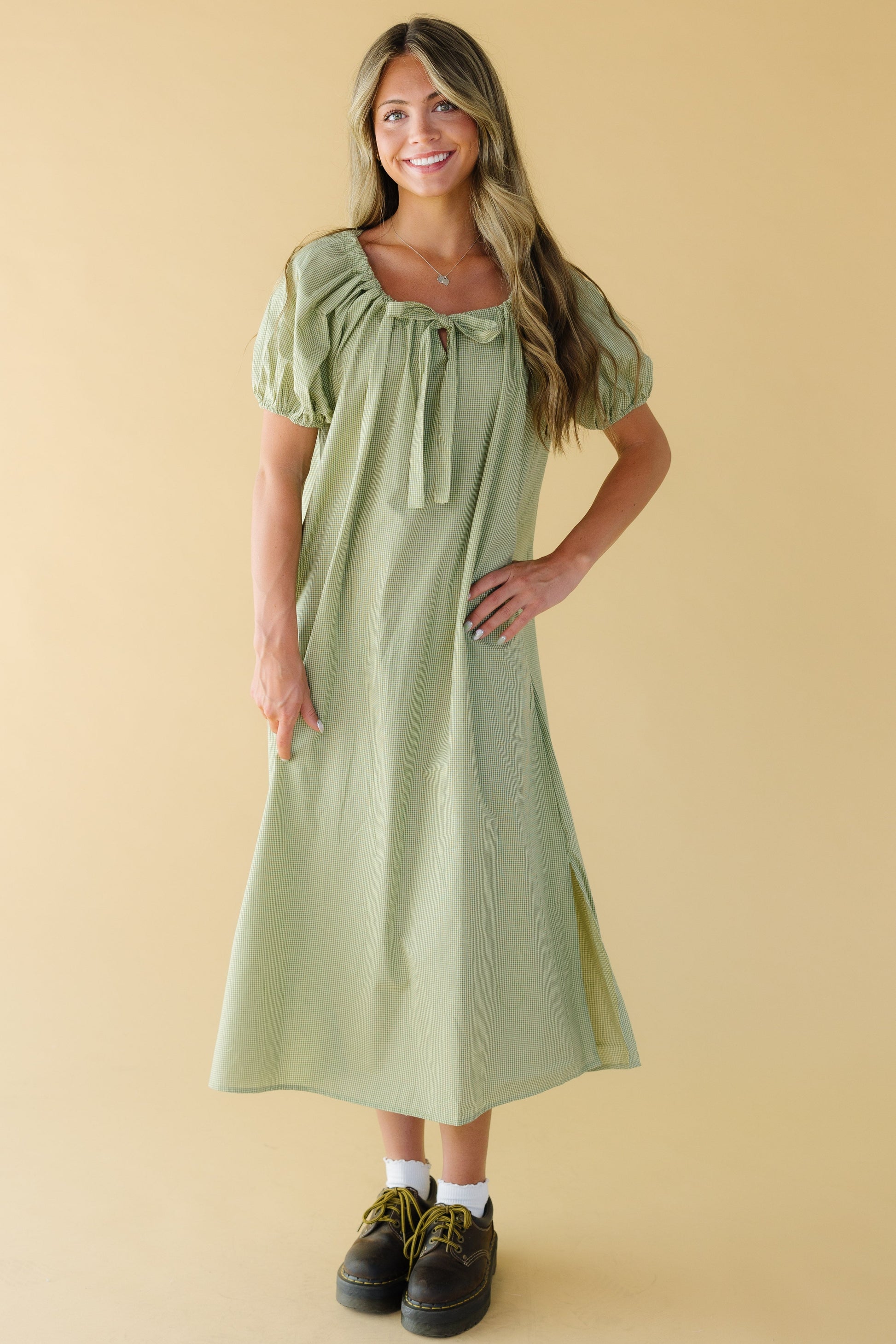 Woman wearing a green gingham midi dress with a square neckline with a same fabric drawstring, and large puff sleeves.