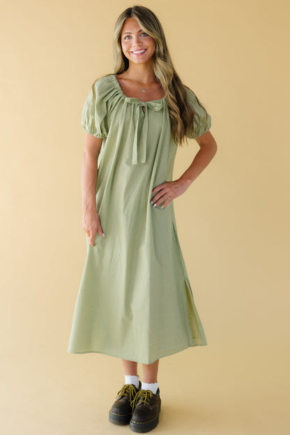Woman wearing a green gingham midi dress with a square neckline with a same fabric drawstring, and large puff sleeves.