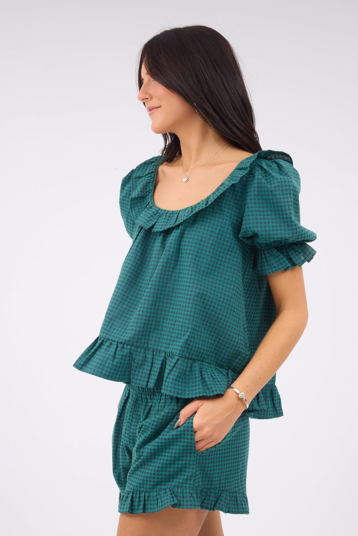 A closer view of a woman wearing a loose fitting green & navy gingham shorts set with short puff sleeves, wide ruffled scoop neckline, ruffled hem.  The model  is albo wearing loose fitting matching shorts with an elastic waistband, side pocketsand a ruffled hem.