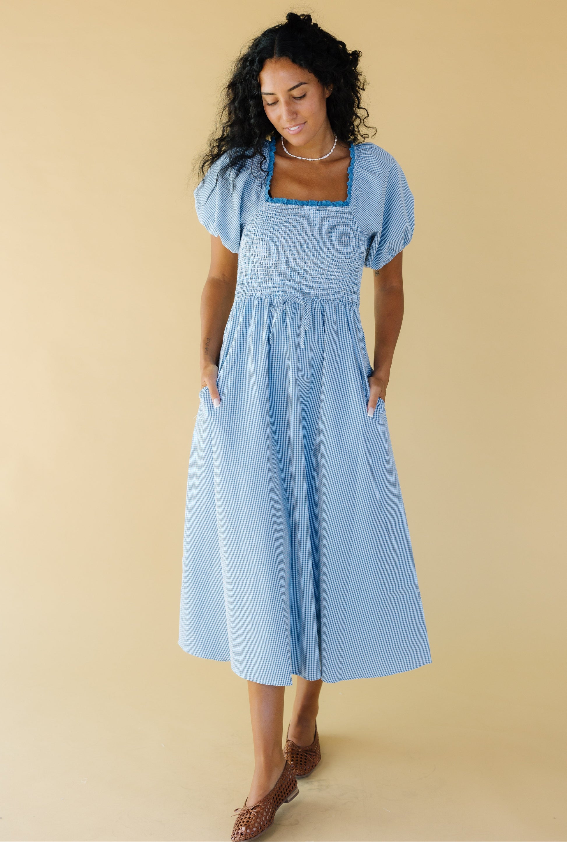 Young woman wearing a modest blue & white gingham midi dress with a lace trimmed square neckline, balloon sleeves, a smocked bodice and side pockets.