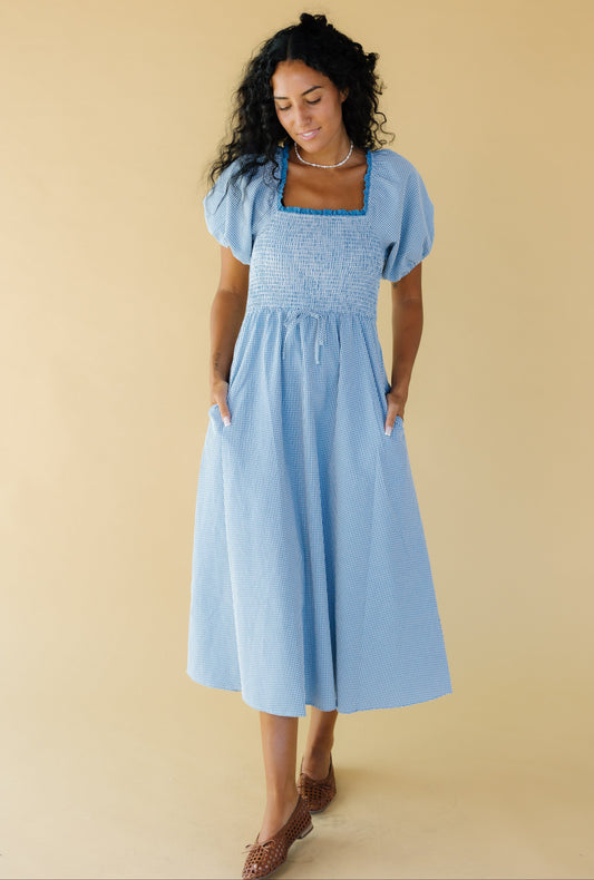 Young woman wearing a modest blue & white gingham midi dress with a lace trimmed square neckline, balloon sleeves, a smocked bodice and side pockets.