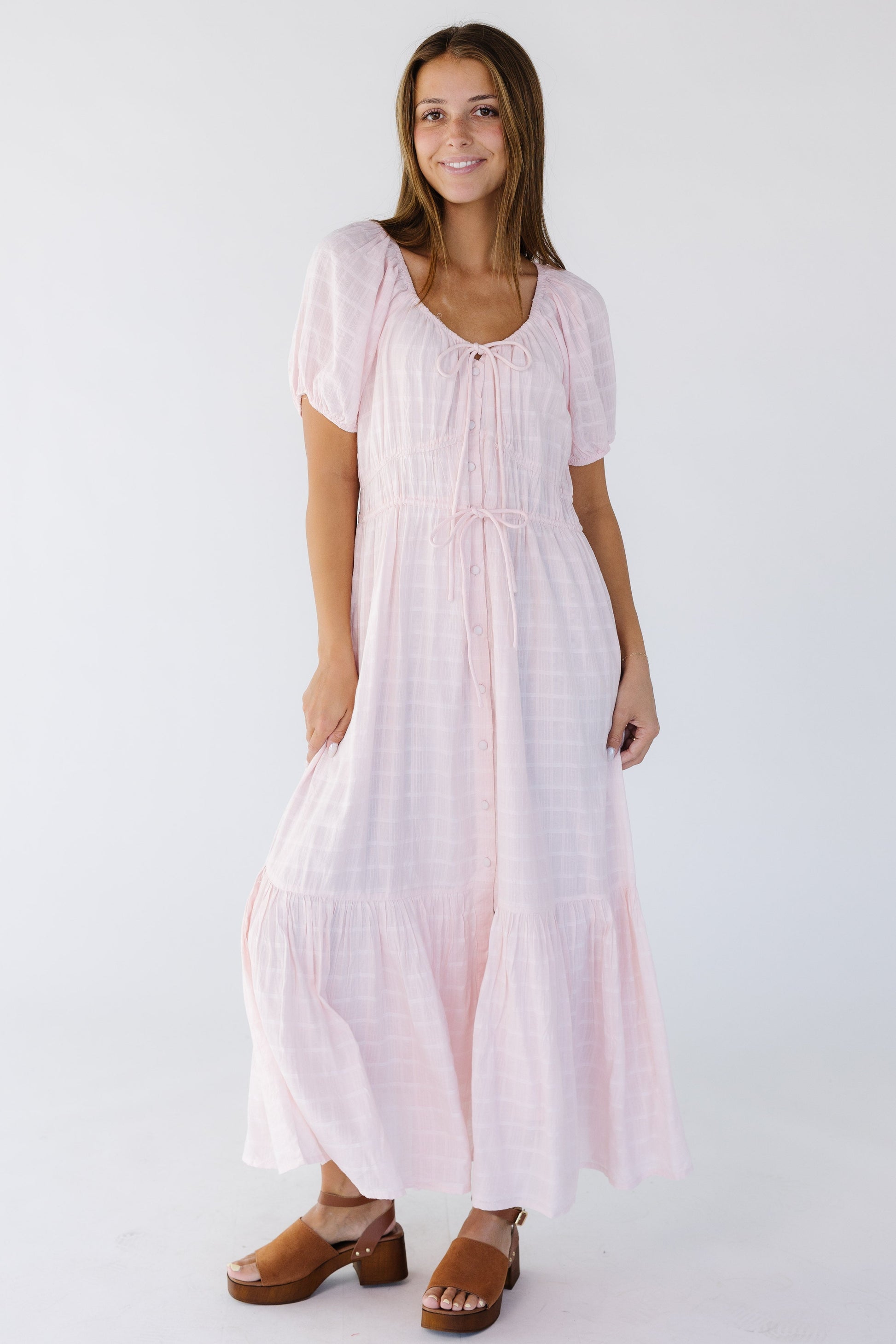 Woman wearing a light pink button down midi-maxi with a drawstring neckline, short puff sleeves and wide bottom ruffle.