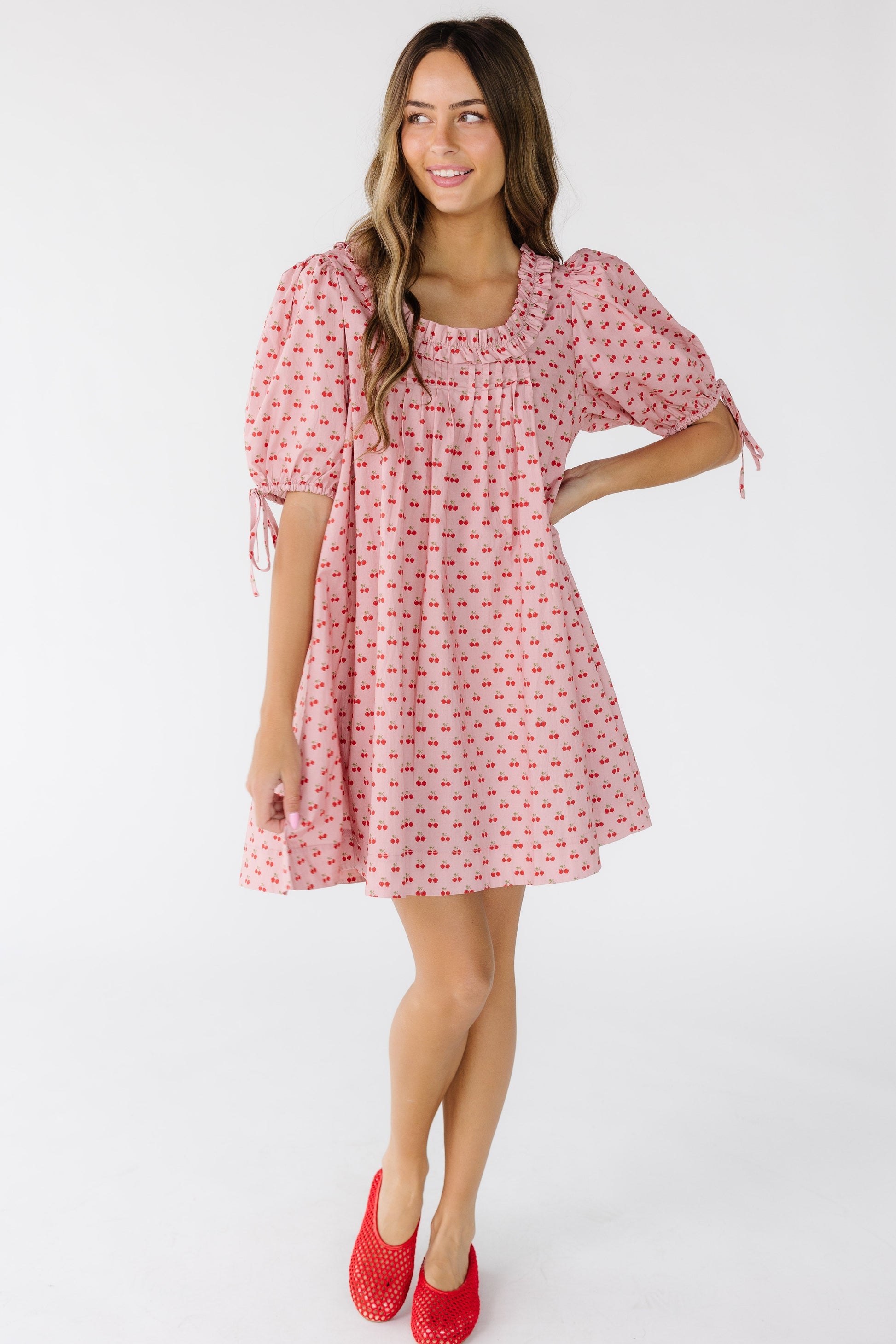 Young woman wearing a pink with cherry print short dress with large balloon sleeves and a ruffled square neckline.