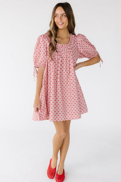 Young woman wearing a pink with cherry print short dress with large balloon sleeves and a ruffled square neckline.