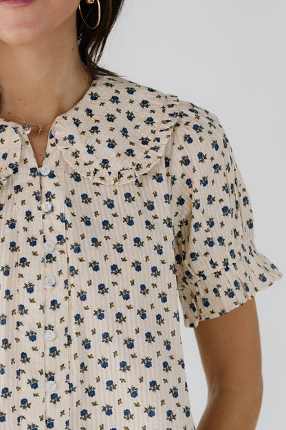 Close view of a woman wearing a cream & blue floral button down blouse with a ruffle edged round collar and short sleeve.