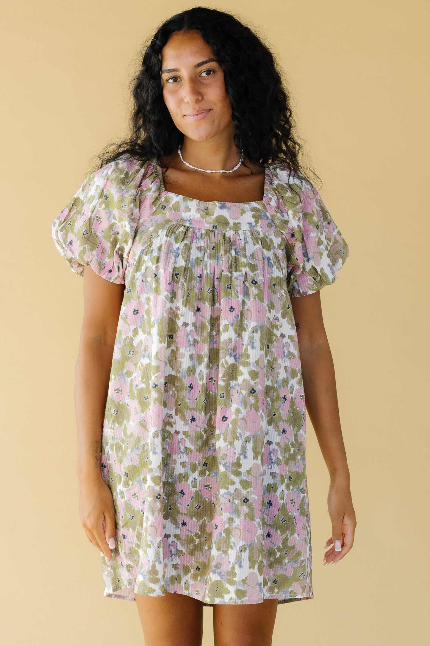 Young woman wearing a short floral dress in pink & green with balloon sleeves and a square neckline.