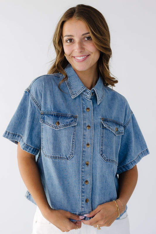 Young woman wearing a modest light denim button down top with a pointed collar, short sleeves and 2 front patch pockets that have button down flaps.