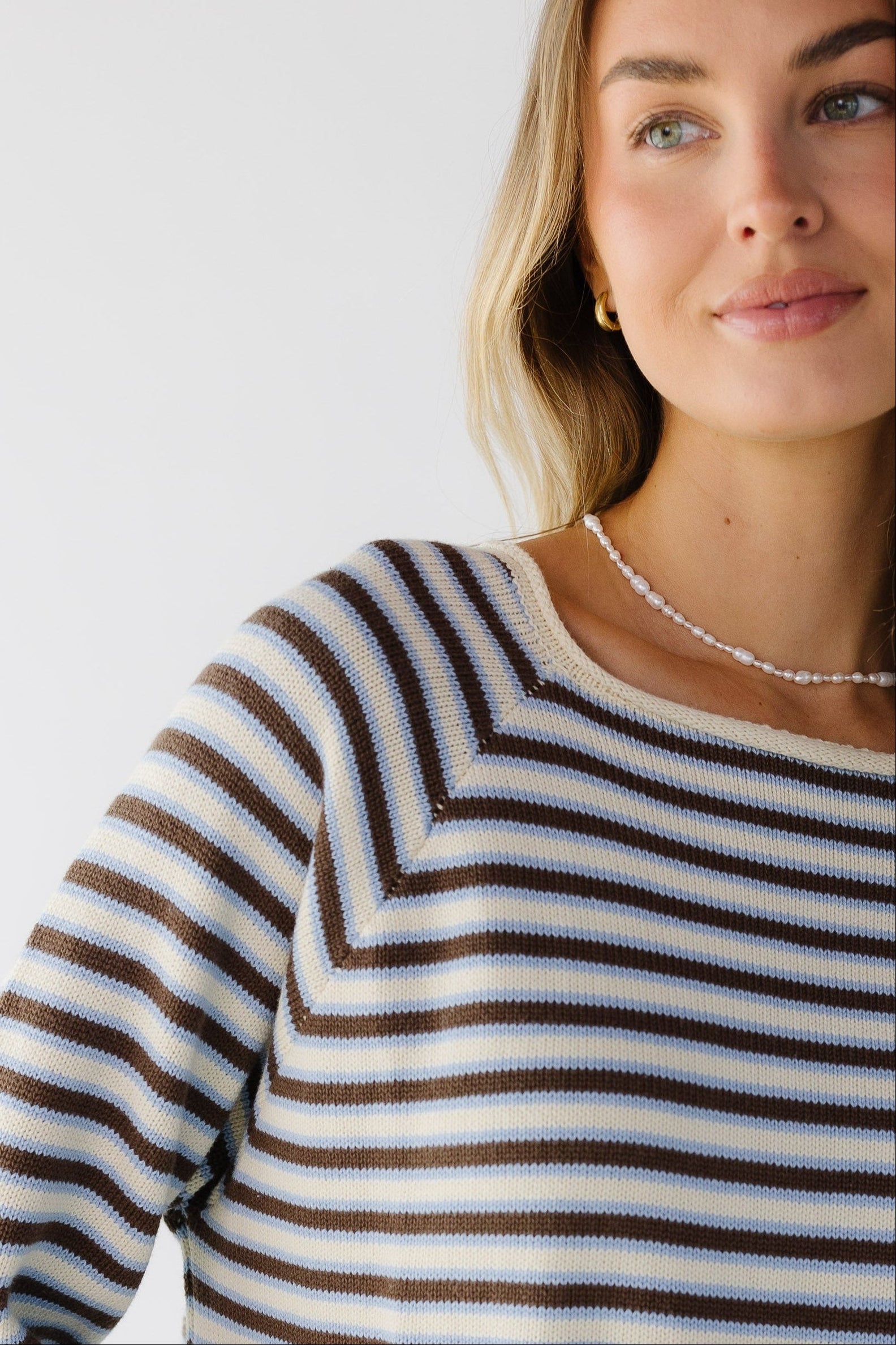 Close view of a woman wearing a cream, blue & brown stripe cotton sweater with a wide rolled neckline.