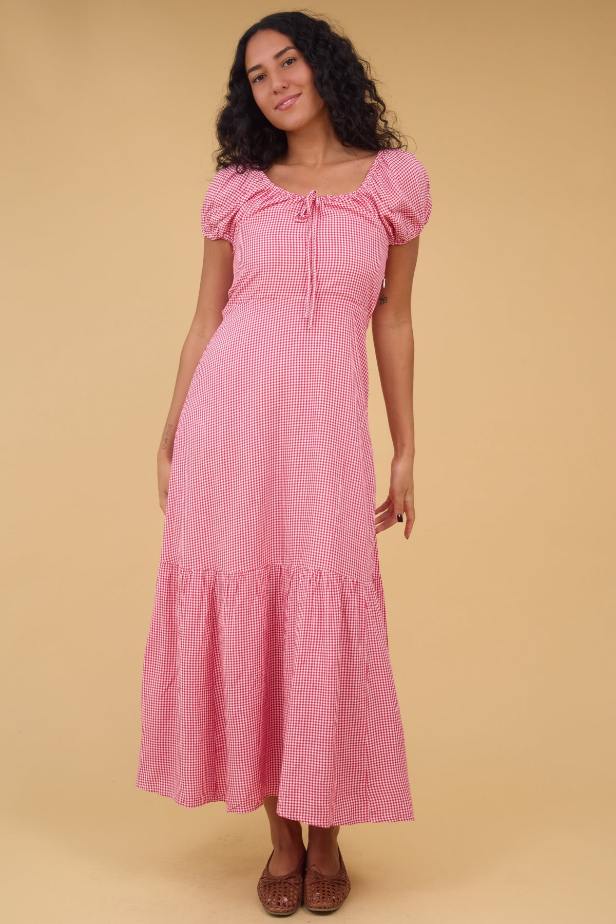 A young woman wearing a pinkgingham midi dress with a scoop drawstring neckline, short puff sleeves and a wide bottom ruffle.