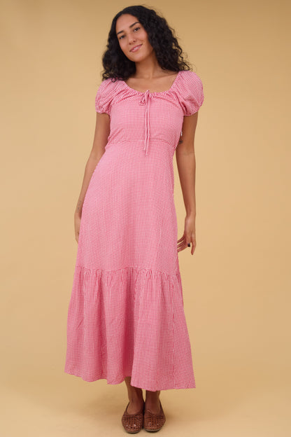 A young woman wearing a pinkgingham midi dress with a scoop drawstring neckline, short puff sleeves and a wide bottom ruffle.