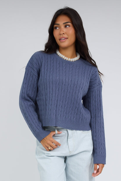 Young woman wearing a modest blue cable knit pullover sweater with a crew neckline and long sleeves.