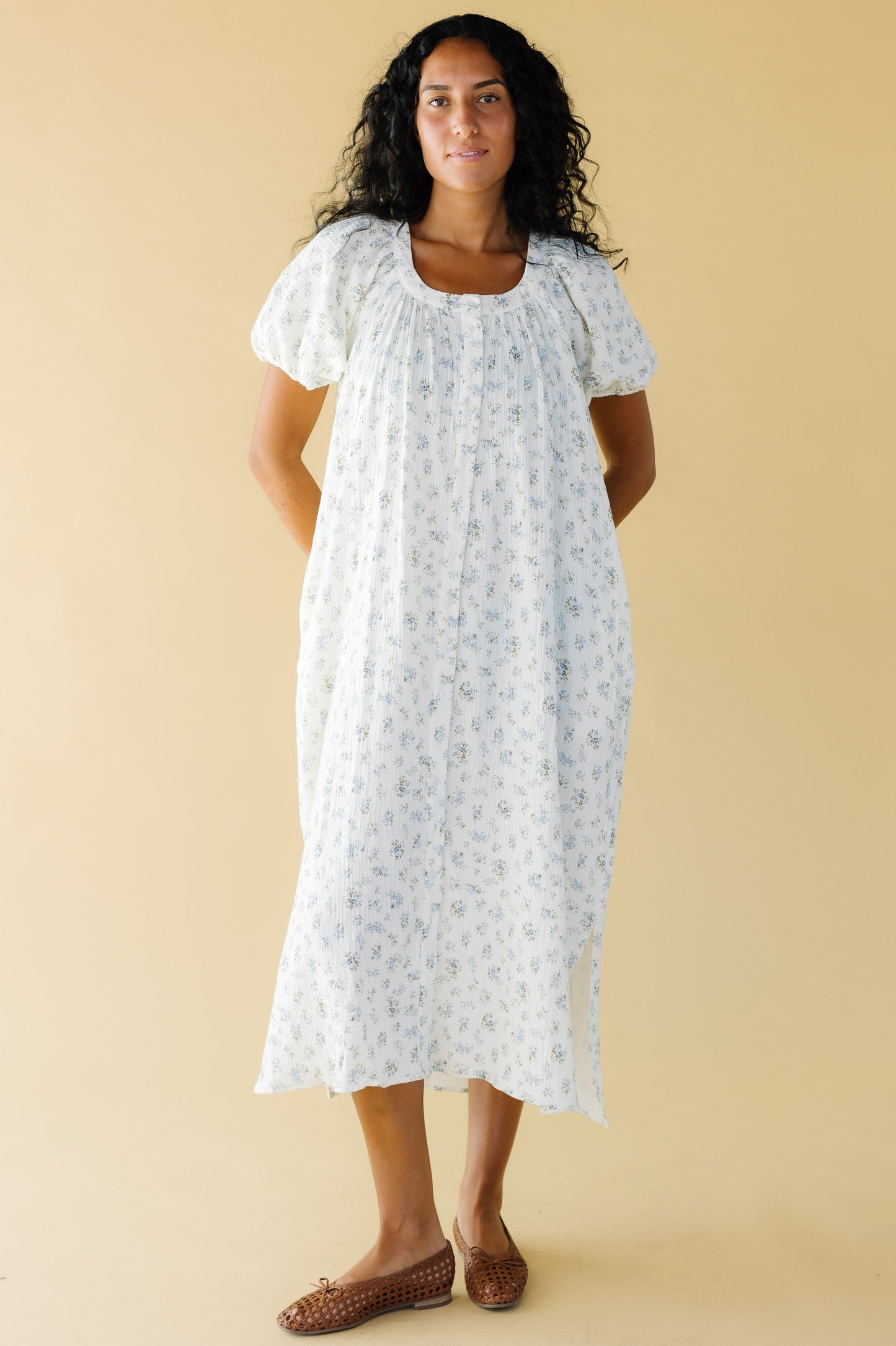 Young woman wearing a modest white & blue floral print midi dress that buttons down the front with balloon sleeves and a scoop neckline.