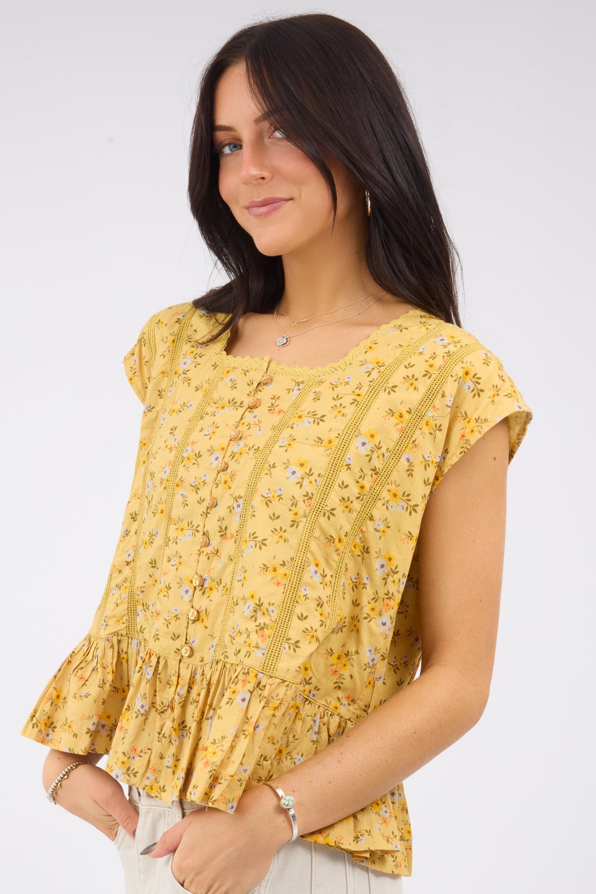 A woman wearing a floral print mustard yellow blouse with cap sleeves, bottom ruffle and lace details.
