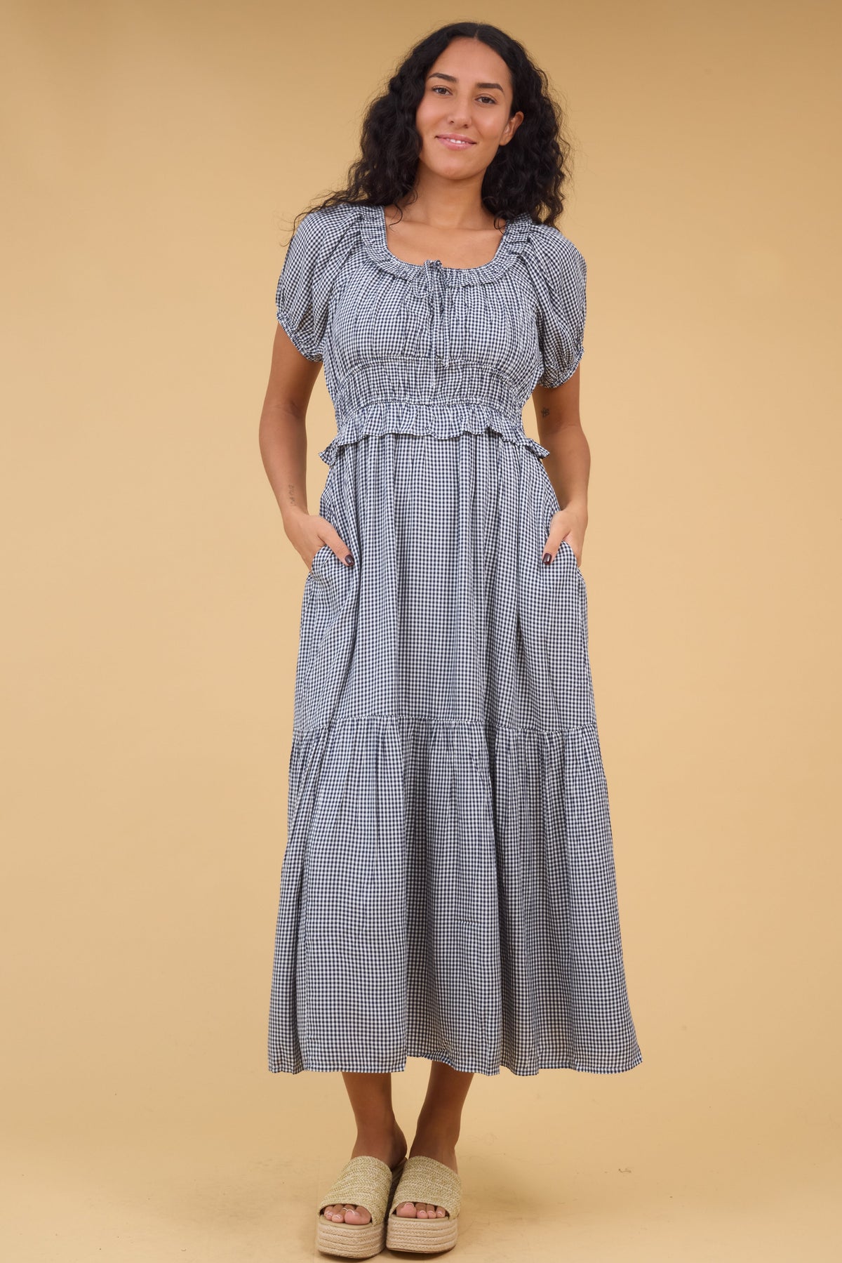 Woman wearing a navy & white gingham midi dress with side pockets, wide bottom ruffle, short puff sleeves and a ruffled scoop neckline.