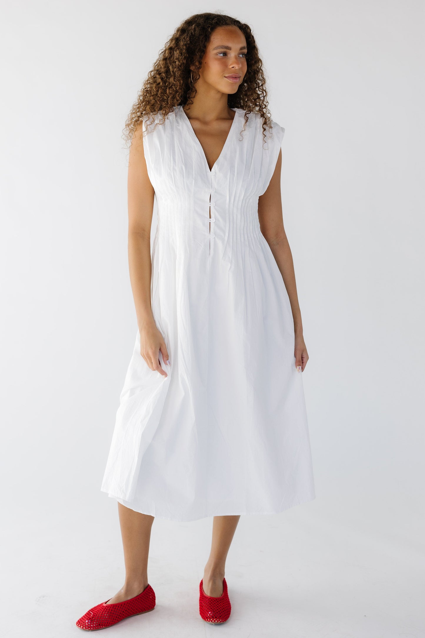 Teenage girl wearing a sleeveless white midi dress with a V-neckline, short button placket at the waist with pleats around the waist.