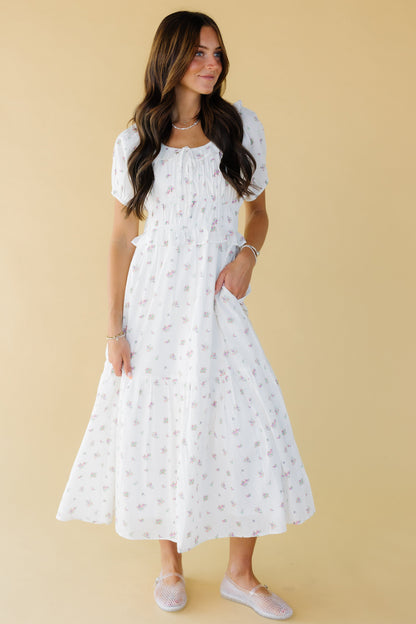 Young woman wearing a modest white floral midi-maxi dress with a scoop neckline, puff sleeves and a wide waistband with a lower ruffle.
