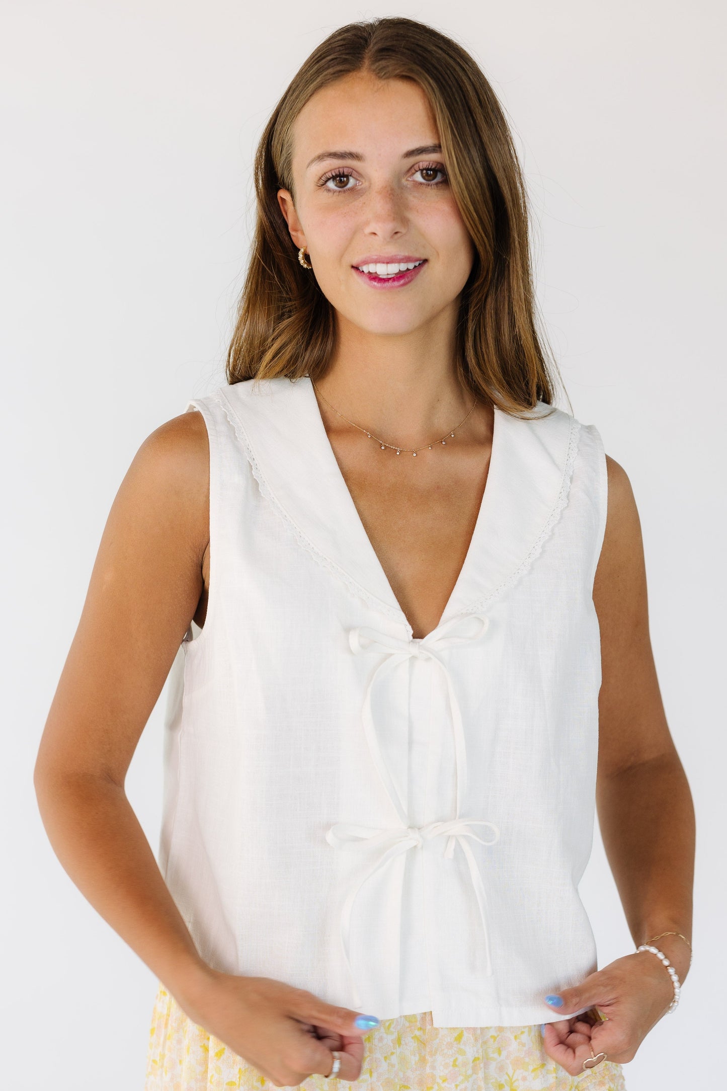 Woman wearing a white sleeveless top with a 2 tie closure and a V-neckline with a lace edge shawl collar.