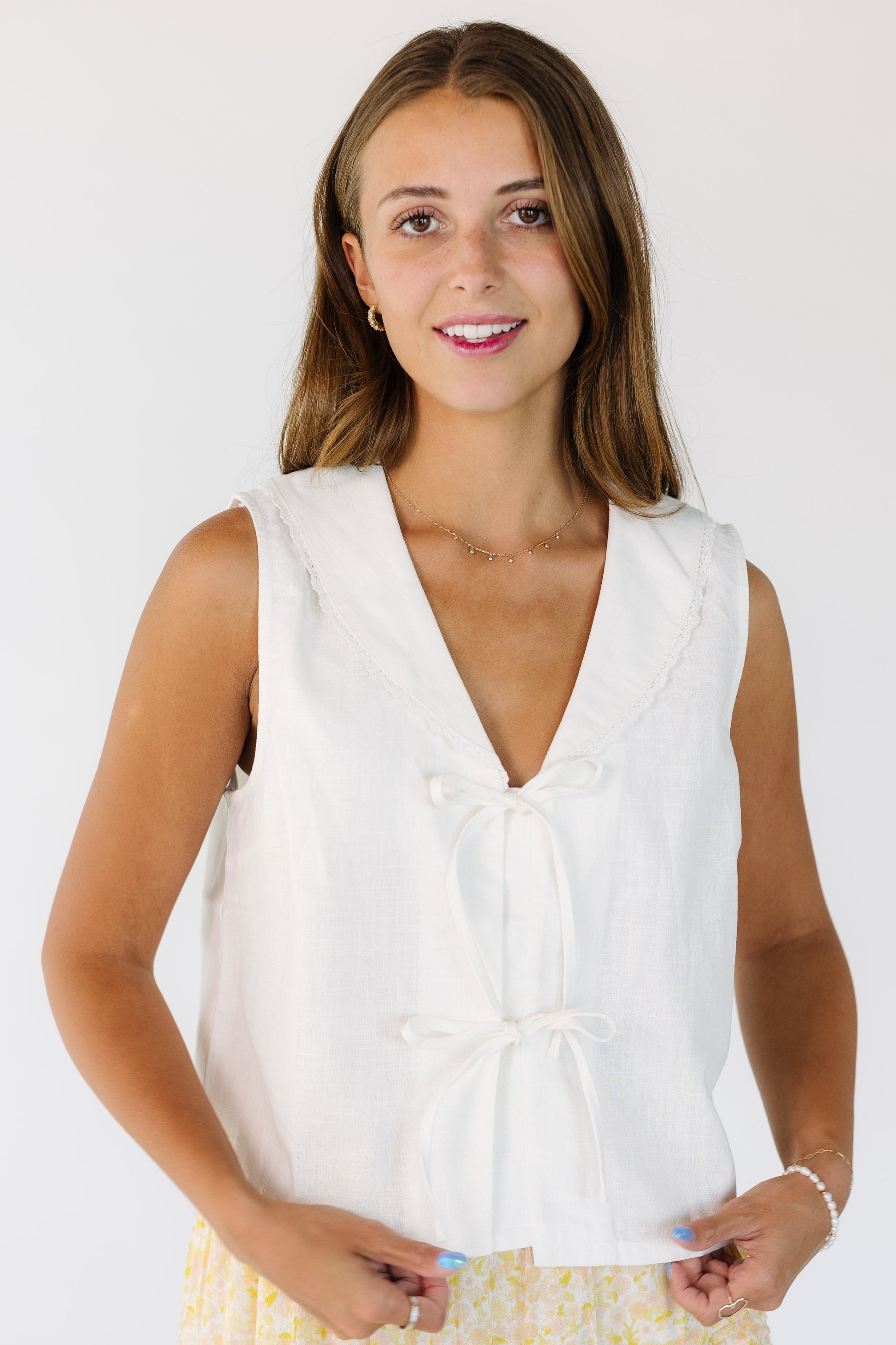 Woman wearing a white sleeveless top with a 2 tie closure and a V-neckline with a lace edge shawl collar.