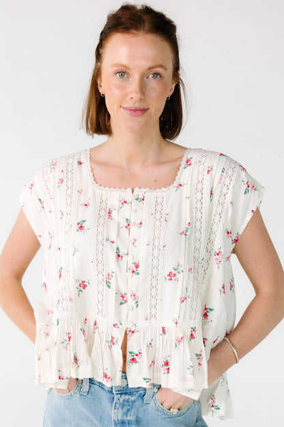 Woman wearing a loose fitting off white and pink floral, button down blouse with a lace trimmed square neckline and a bottom ruffle.