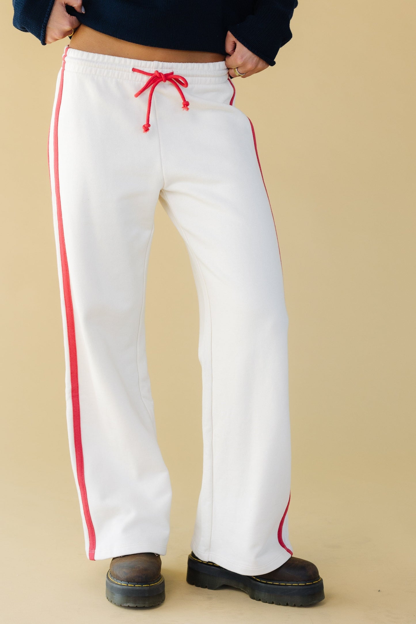 Young woman wearing white lounge pants with an elastic & drawstring waist, double stripes down the side and a wide cut leg.