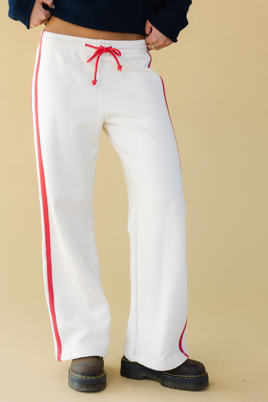 Young woman wearing white lounge pants with an elastic & drawstring waist, double stripes down the side and a wide cut leg.