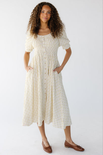 Young woman wearing a cream floral, button down midi dress with a wide scoop neckline with a wide smocked waist and side pockets.