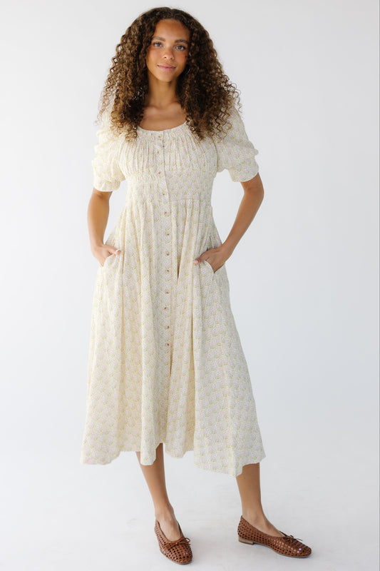 Young woman wearing a cream floral, button down midi dress with a wide scoop neckline with a wide smocked waist and side pockets.